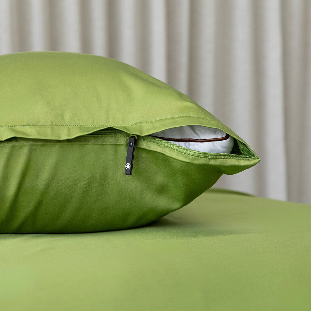 Green pillow with zipper on a bed against a neutral curtain background.