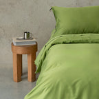 Green bedding set on a bed with a wooden side table and concrete wall background.