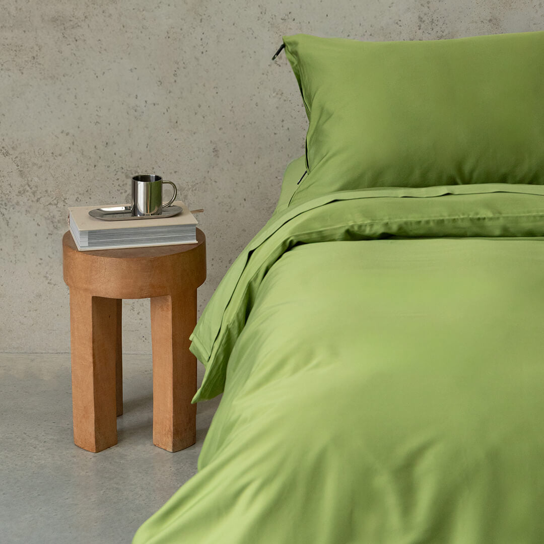 Green bedding set on a bed with a wooden side table and concrete wall background.
