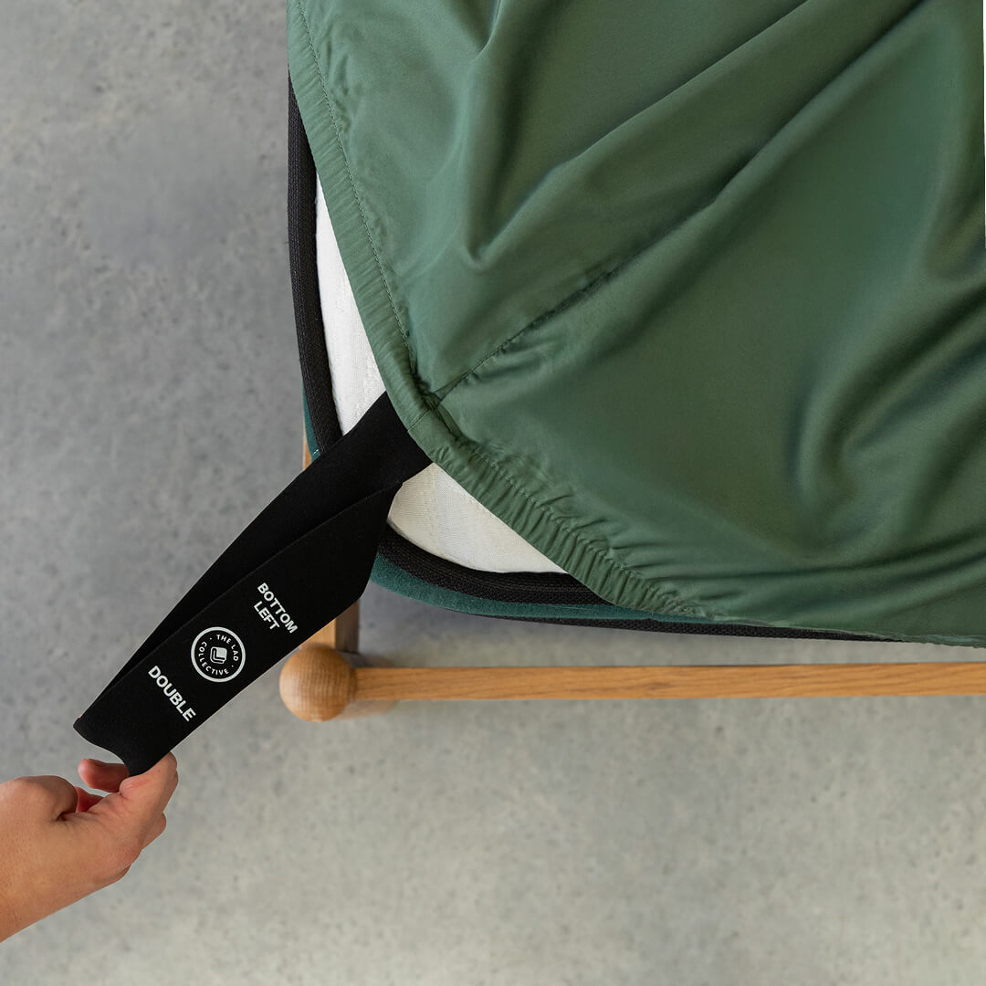 Person pulling labelled corner loop strap on green fitted sheet.