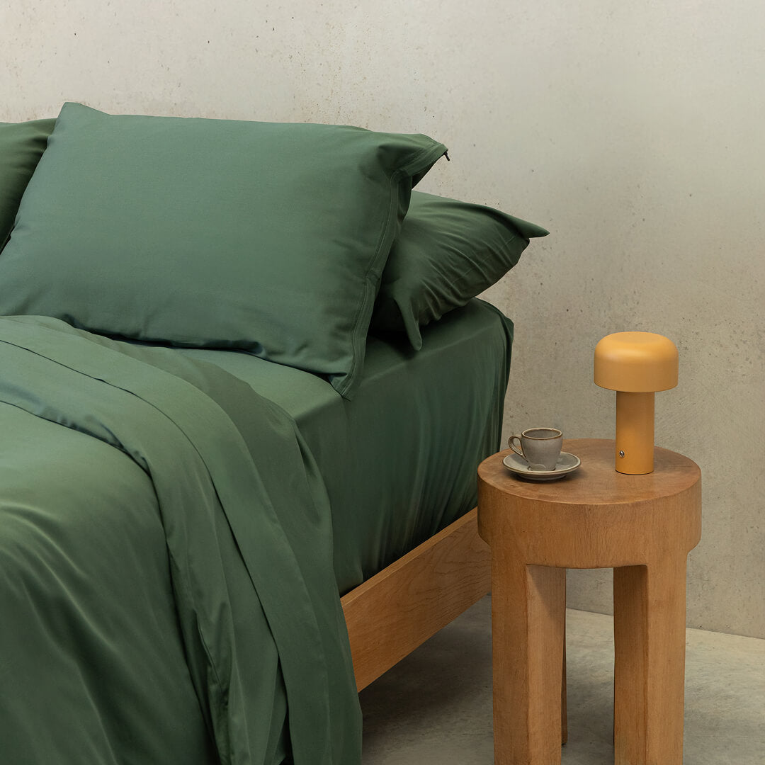 Angled shot of a dark green quilt cover on a bed, with a bedside table to the right and an orange lamp.