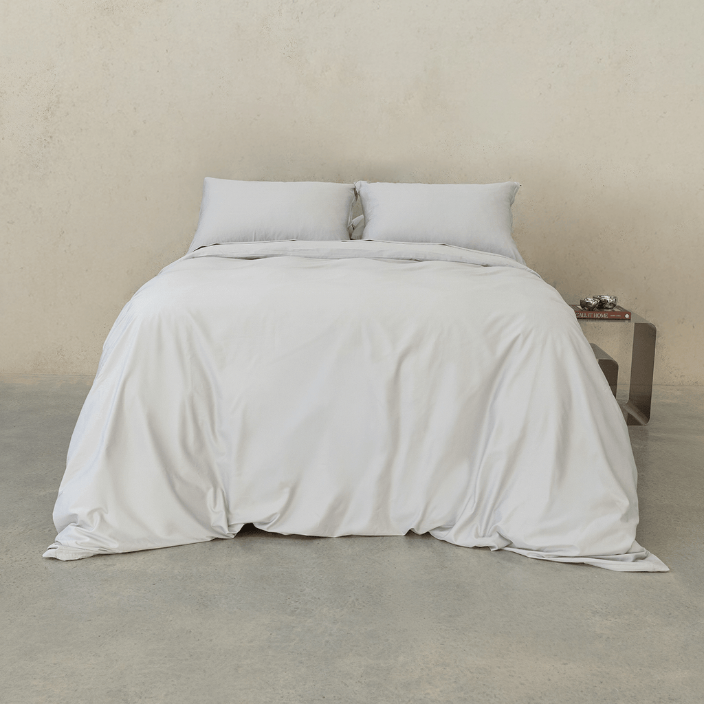 Front view of a bed dressed in soft gray bedding with two matching pillows and a gray quilt cover draped to the floor against a neutral concrete wall.