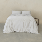 Front view of a bed dressed in soft gray bedding with two matching pillows and a gray quilt cover draped to the floor against a neutral concrete wall.