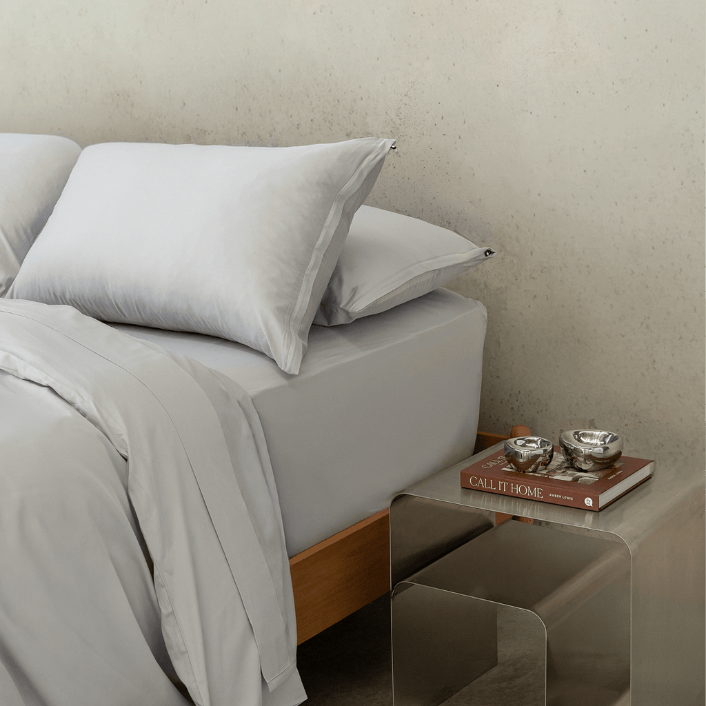 Close-up of layered gray pillows and bedding beside a metallic bedside table with stacked books and decorative silver bowls.