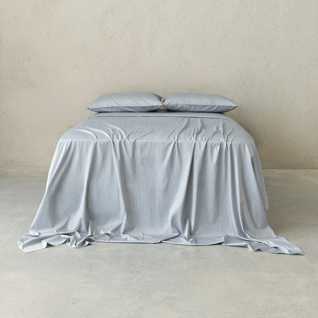 Front view of a bed dressed in gray and white pinstripe bedding with two matching pillows and a softly draped sheet against a neutral concrete wall.