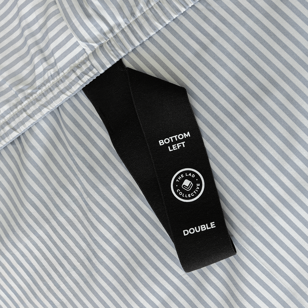 Close-up of a black fitted-sheet corner strap labelled “Bottom Left” and “Double” against gray and white striped fabric.