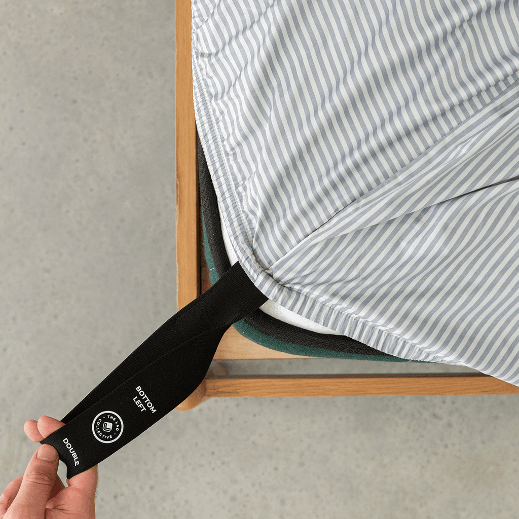 A hand pulls the labelled black corner strap of a gray and white striped fitted sheet around the bottom-left corner of a double mattress.