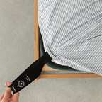 A hand pulls the labelled black corner strap of a gray and white striped fitted sheet around the bottom-left corner of a double mattress.