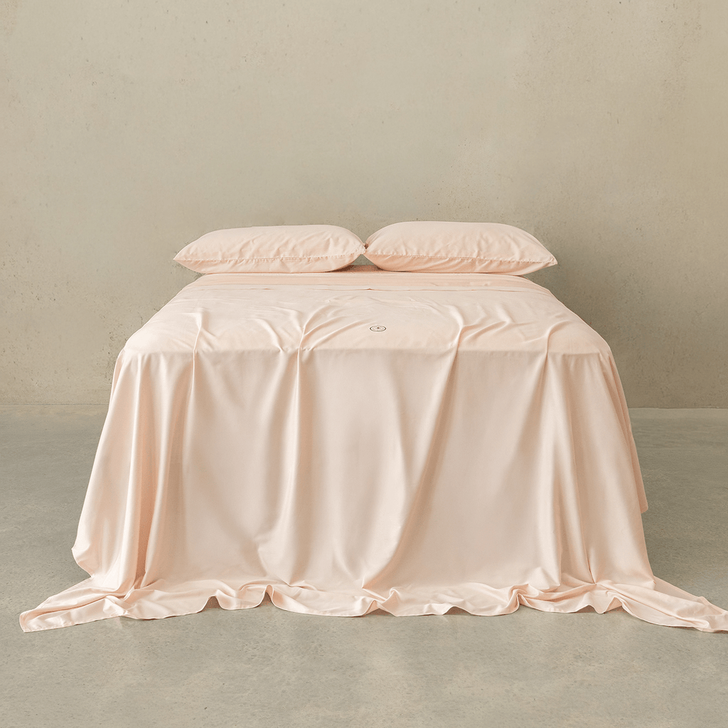 Soft blush pink fitted sheet set styled on a neatly made bed with matching pillowcases, featuring a smooth drape, elegant finish, and calming bedroom aesthetic.