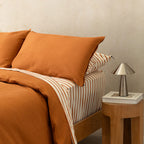 Bed with orange bedding and striped sheets, next to a wooden nightstand with a lamp.