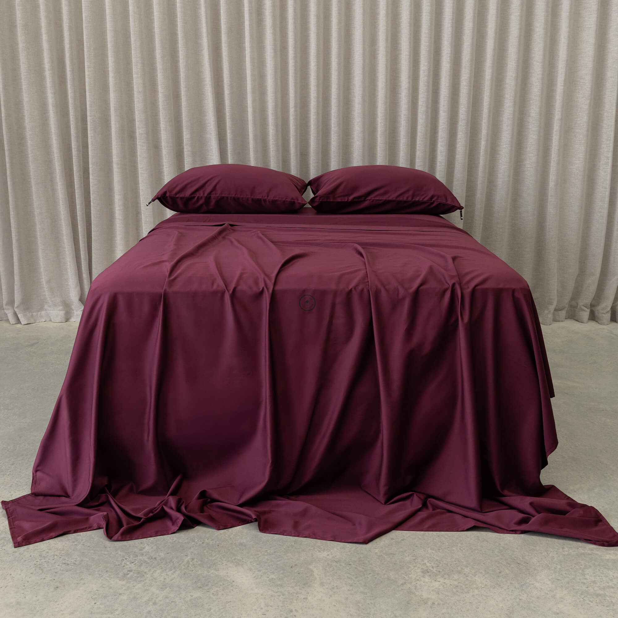 Maroon bedspread and pillows on a bed with beige curtains in the background