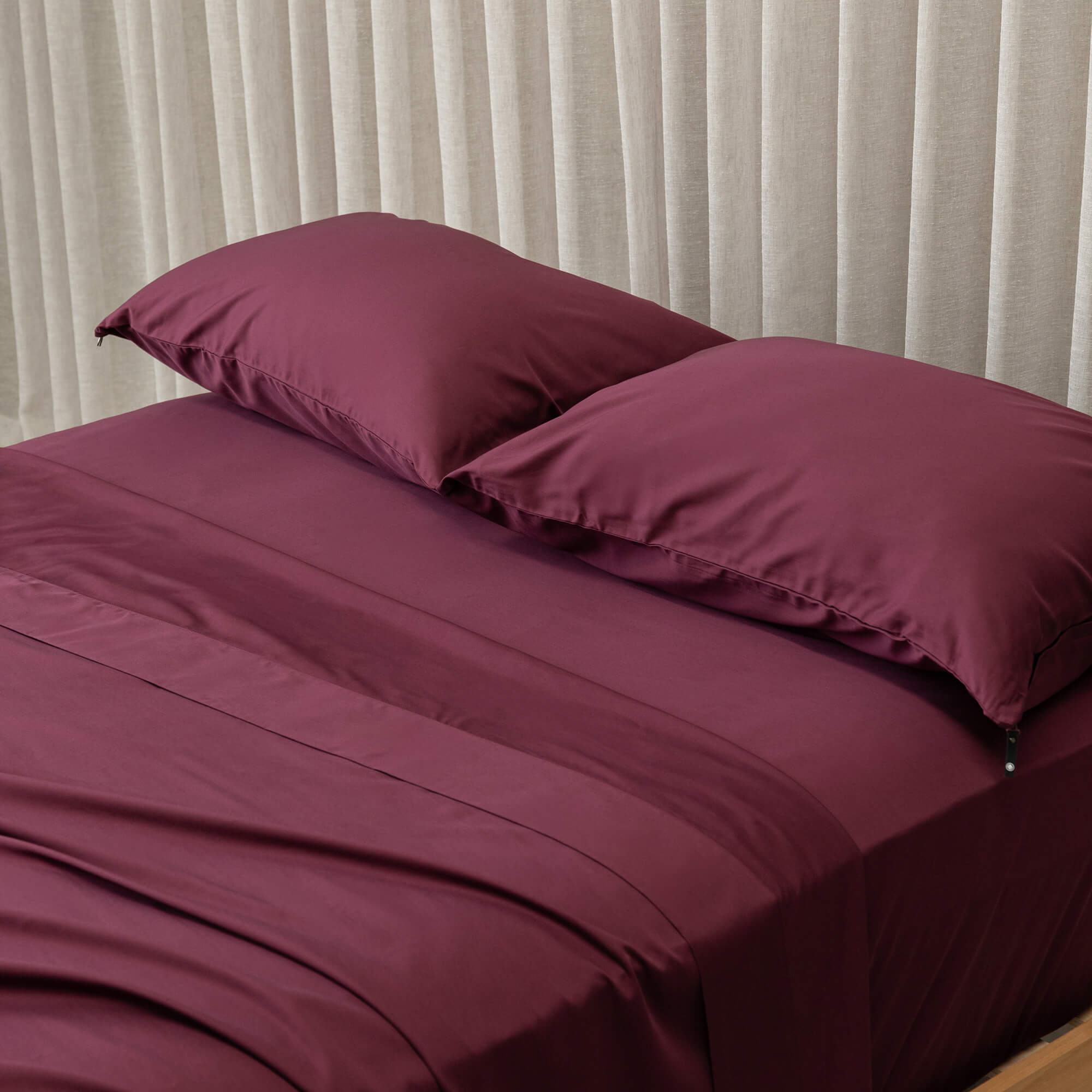Maroon bedspread and pillows on a bed with beige curtains in the background