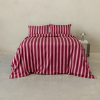 Front view of a bed styled with a burgundy quilt cover and matching pillowcases featuring vertical pale pink stripes, beside a metallic bedside table and lamp.