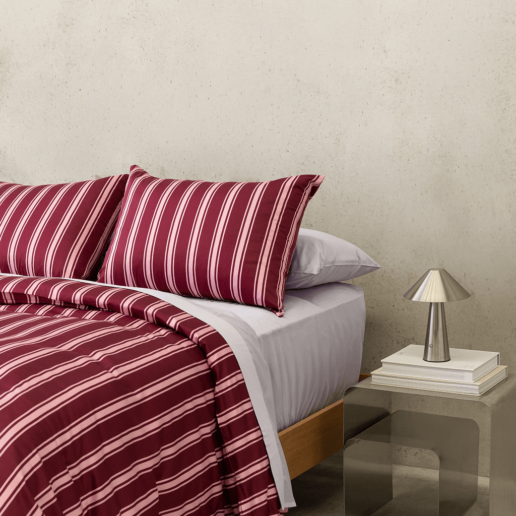 Close-up of burgundy and pale pink striped bedding layered over light-coloured sheets, with a metallic bedside lamp and stacked books.