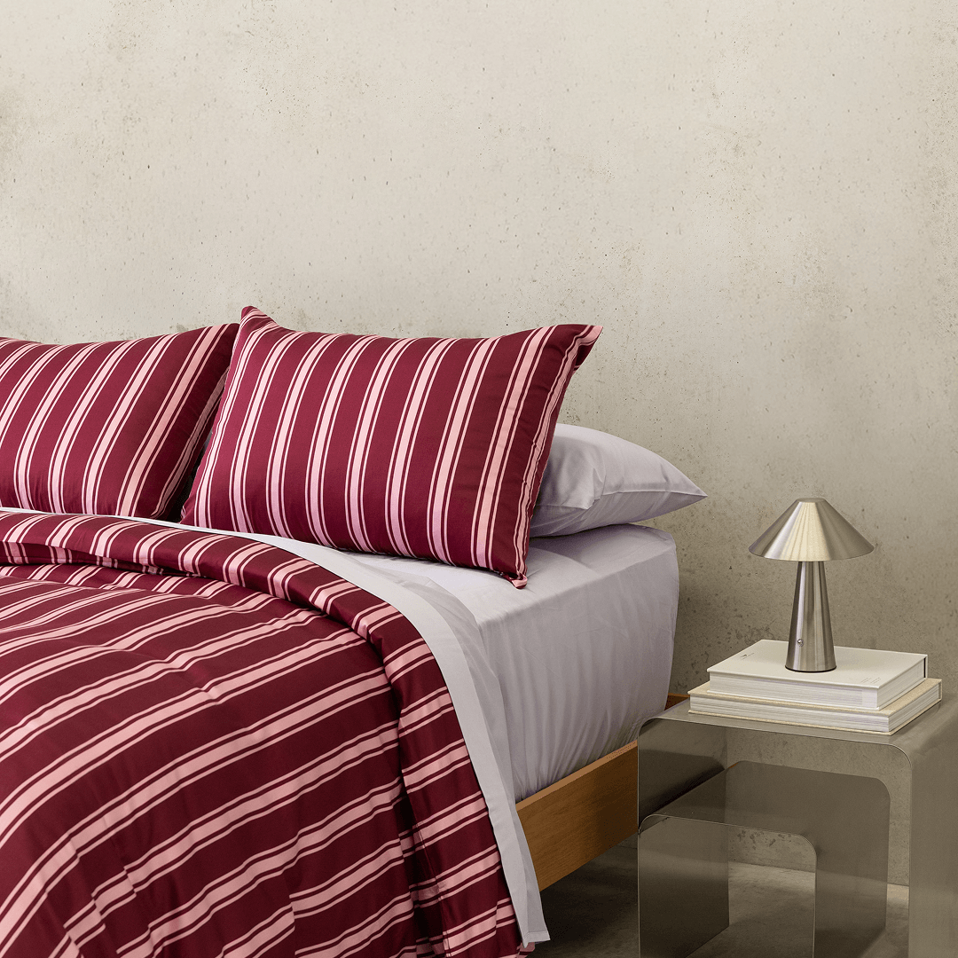 Close-up of burgundy and pale pink striped bedding layered over light-coloured sheets, with a metallic bedside lamp and stacked books.