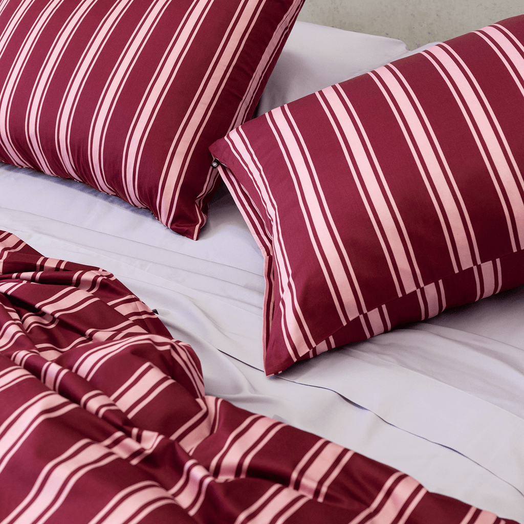 Two burgundy and pale pink striped pillows arranged on light-coloured sheets with the matching quilt cover gathered in front.