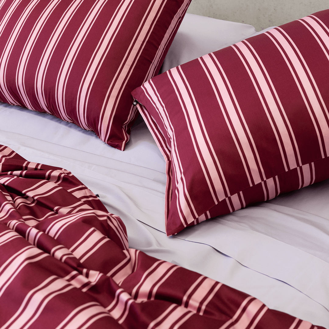 Two burgundy and pale pink striped pillows arranged on light-coloured sheets with the matching quilt cover gathered in front.