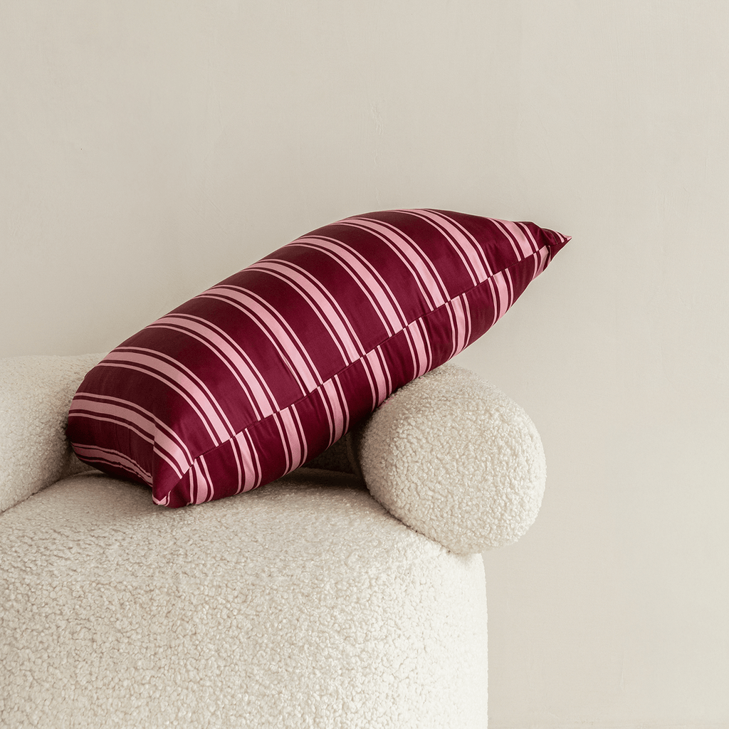 A burgundy and pale pink striped pillow resting diagonally on a cream textured armchair against a neutral wall.
