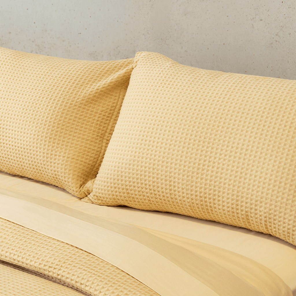 Close-up of two yellow waffle-weave pillowcases leaning against a smooth yellow flat sheet on a bed