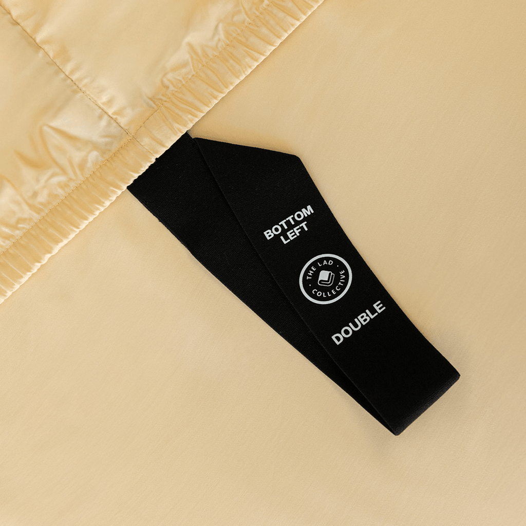 Close-up of a black corner elastic strap on a pale yellow fitted sheet printed with 'Bottom Left' position label