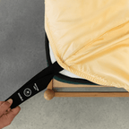 Hand pulling a black elastic corner band labeled 'Bottom Left' while securing a pale yellow fitted sheet onto a mattress corner