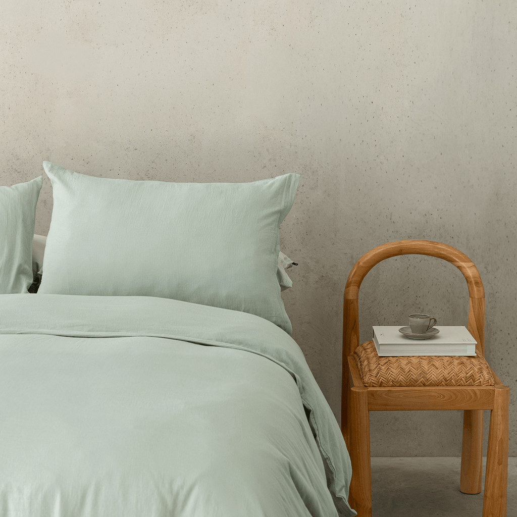 Close-up bedroom styling shot featuring sage green bedding paired with a timber chair, emphasizing the calming color and relaxed texture.