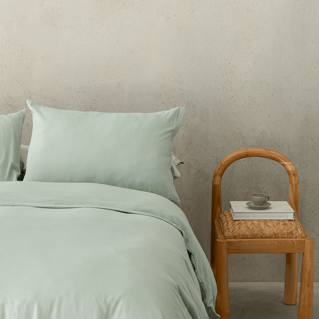 Close-up bedroom styling shot featuring sage green bedding paired with a timber chair, emphasizing the calming color and relaxed texture.