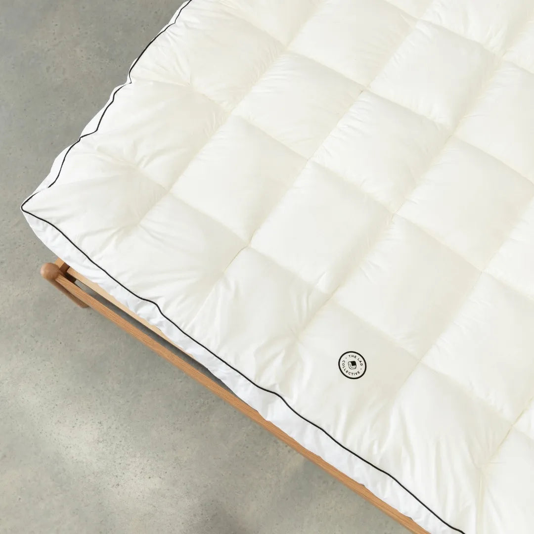 White mattress topper with black trim on a wooden bed frame against a neutral background.