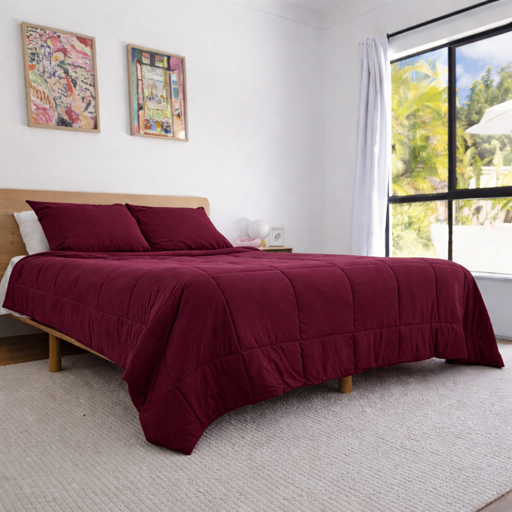 Bed with burgundy comforter and pillows in a bright bedroom with large windows.