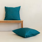 Two teal European pillowcases against a neutral background. One on e timber bench, one on the floor.