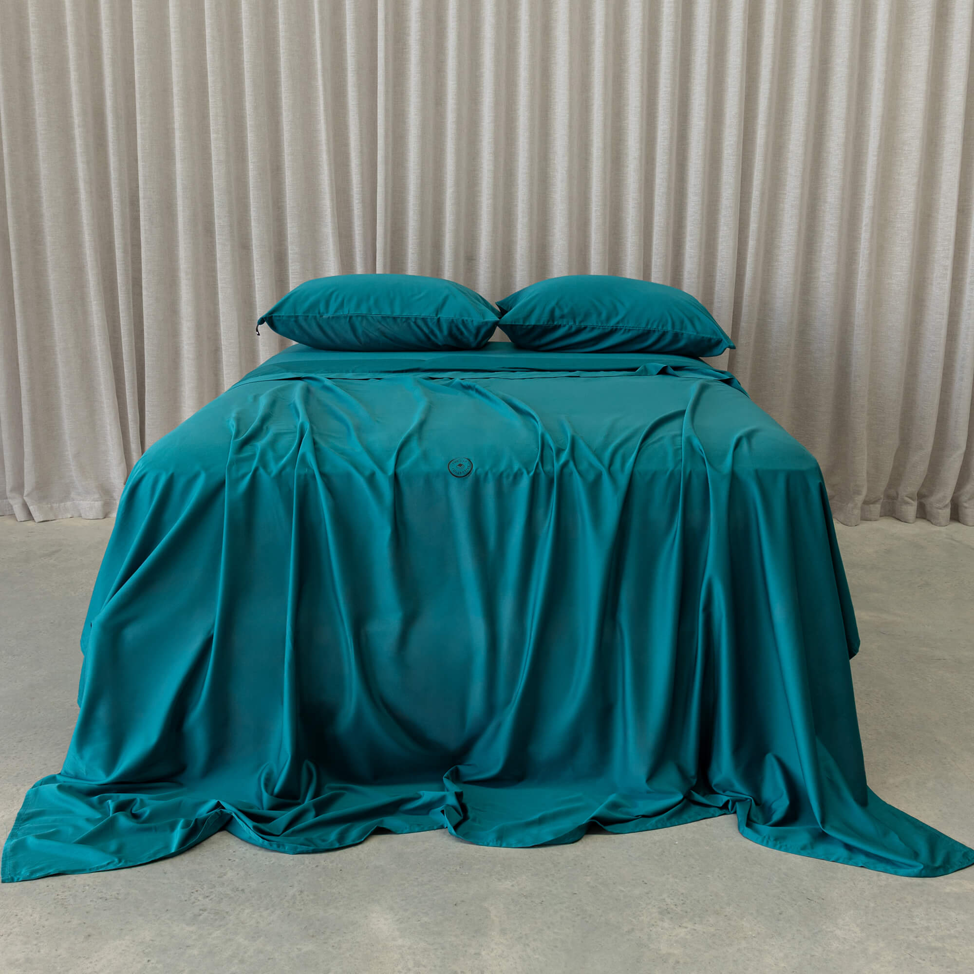Teal bedspread and pillows on a bed with beige curtains in the background