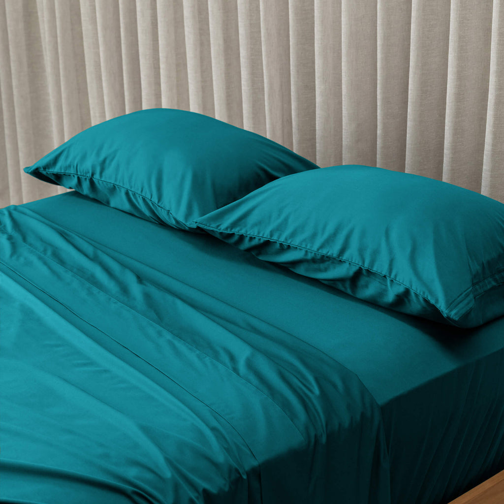 Teal bedspread and pillows on a bed with beige curtains in the background