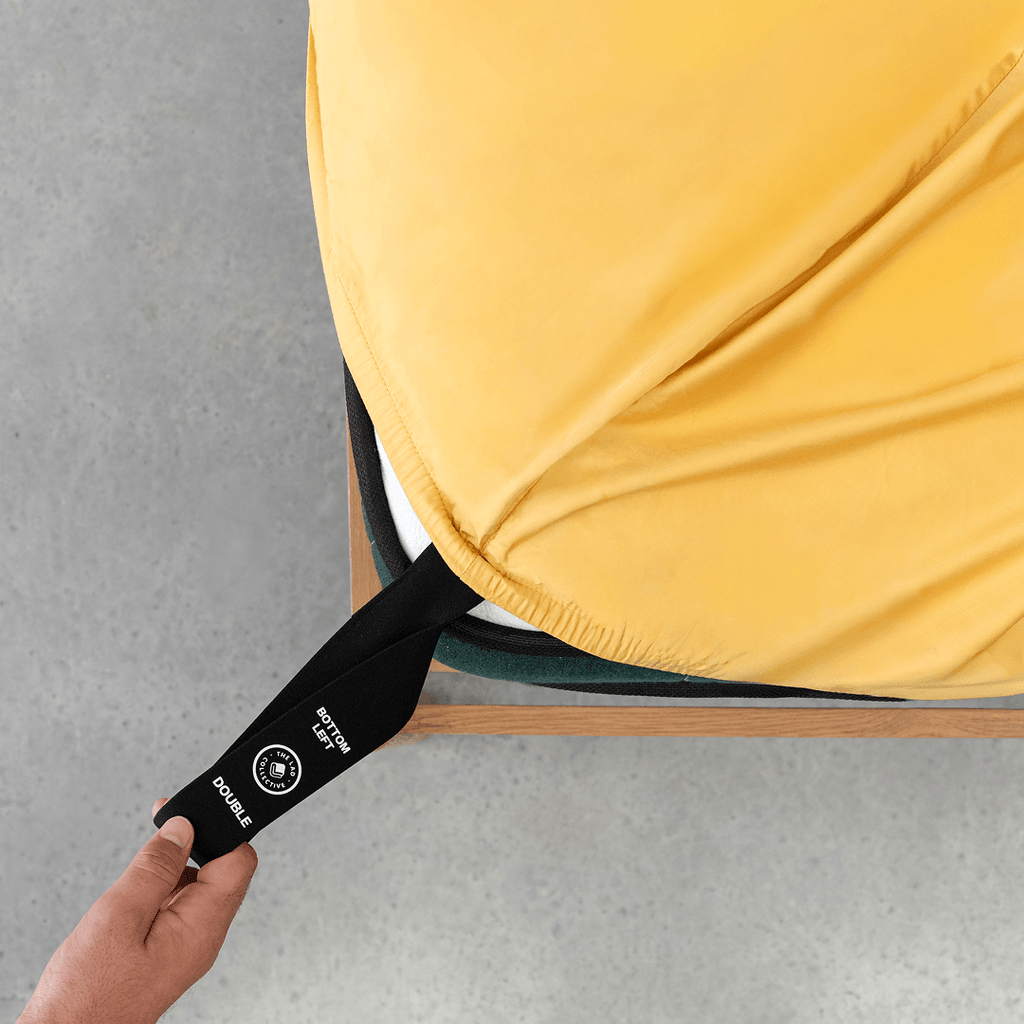 Hand pulling a black elastic corner band labeled 'Bottom Left' while securing a golden yellow fitted sheet onto a mattress corner