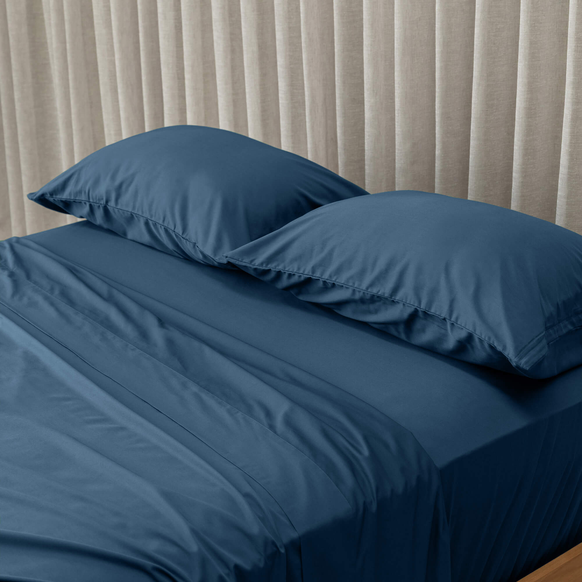 Blue bed sheets and pillows on a bed with beige curtains in the background