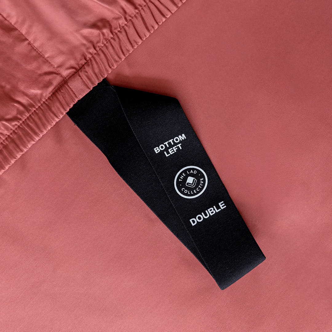 Close-up of a black corner elastic strap on a dusty rose pink fitted sheet printed with 'Bottom Left' position label