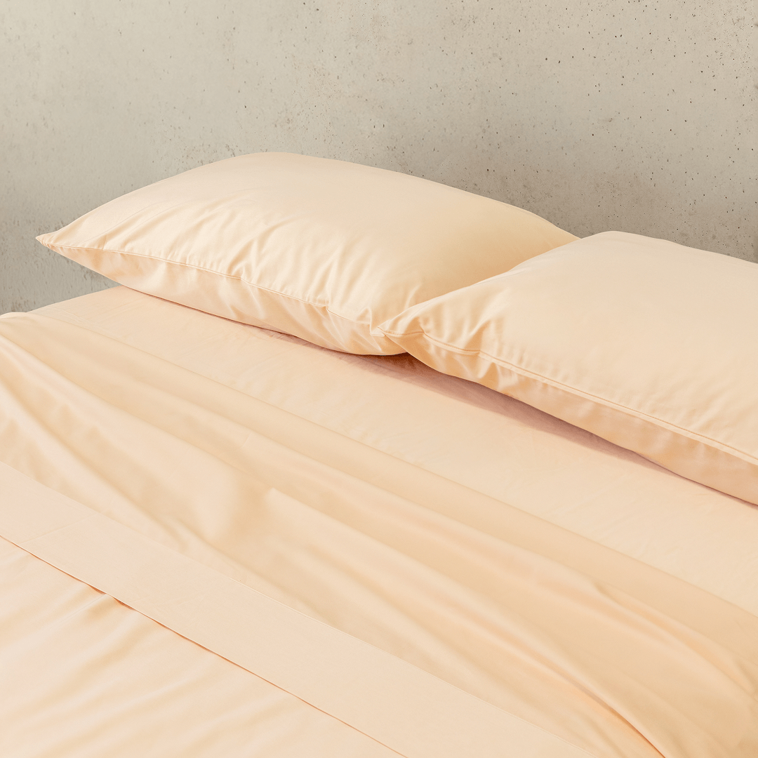 Angle view of two peach pillowcases and a matching flat sheet neatly arranged on a bed