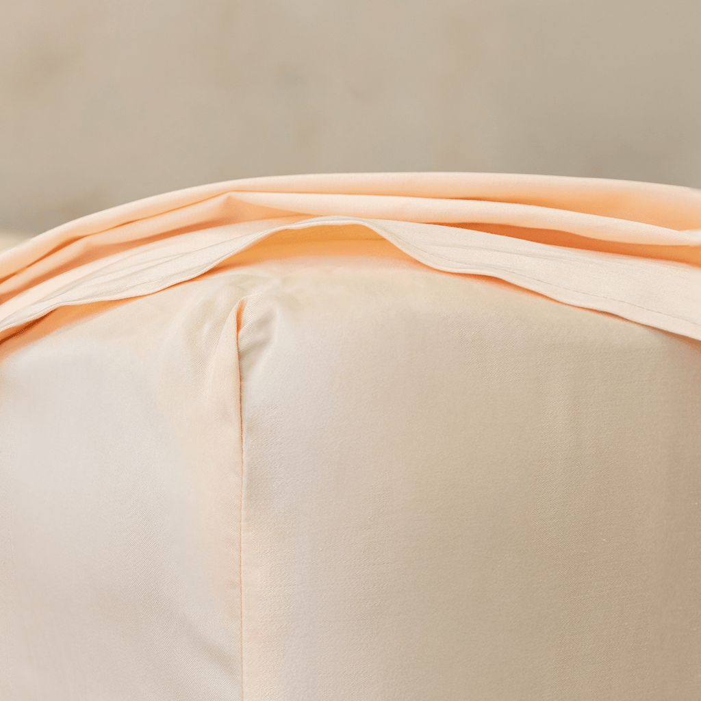 Close-up corner detail of a peach fitted sheet and top flat sheet showing smooth fabric texture and seam stitching