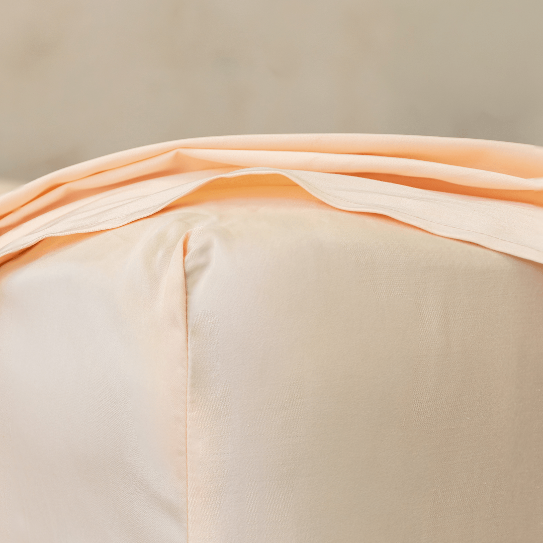 Close-up corner detail of a peach fitted sheet and top flat sheet showing smooth fabric texture and seam stitching