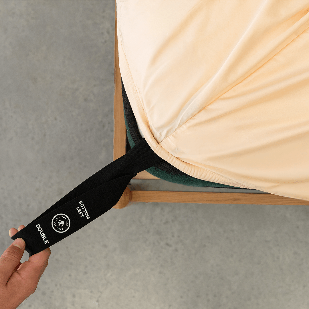 Hand pulling a black elastic corner band labeled 'Bottom Left' while securing a peach fitted sheet onto a mattress corner