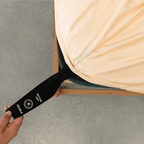Hand pulling a black elastic corner band labeled 'Bottom Left' while securing a peach fitted sheet onto a mattress corner
