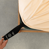 Hand pulling a black elastic corner band labeled 'Bottom Left' while securing a peach fitted sheet onto a mattress corner
