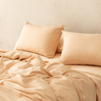 Angle view of two peach pillowcases resting on a matching flat sheet and rumpled quilt cover on a styled bed