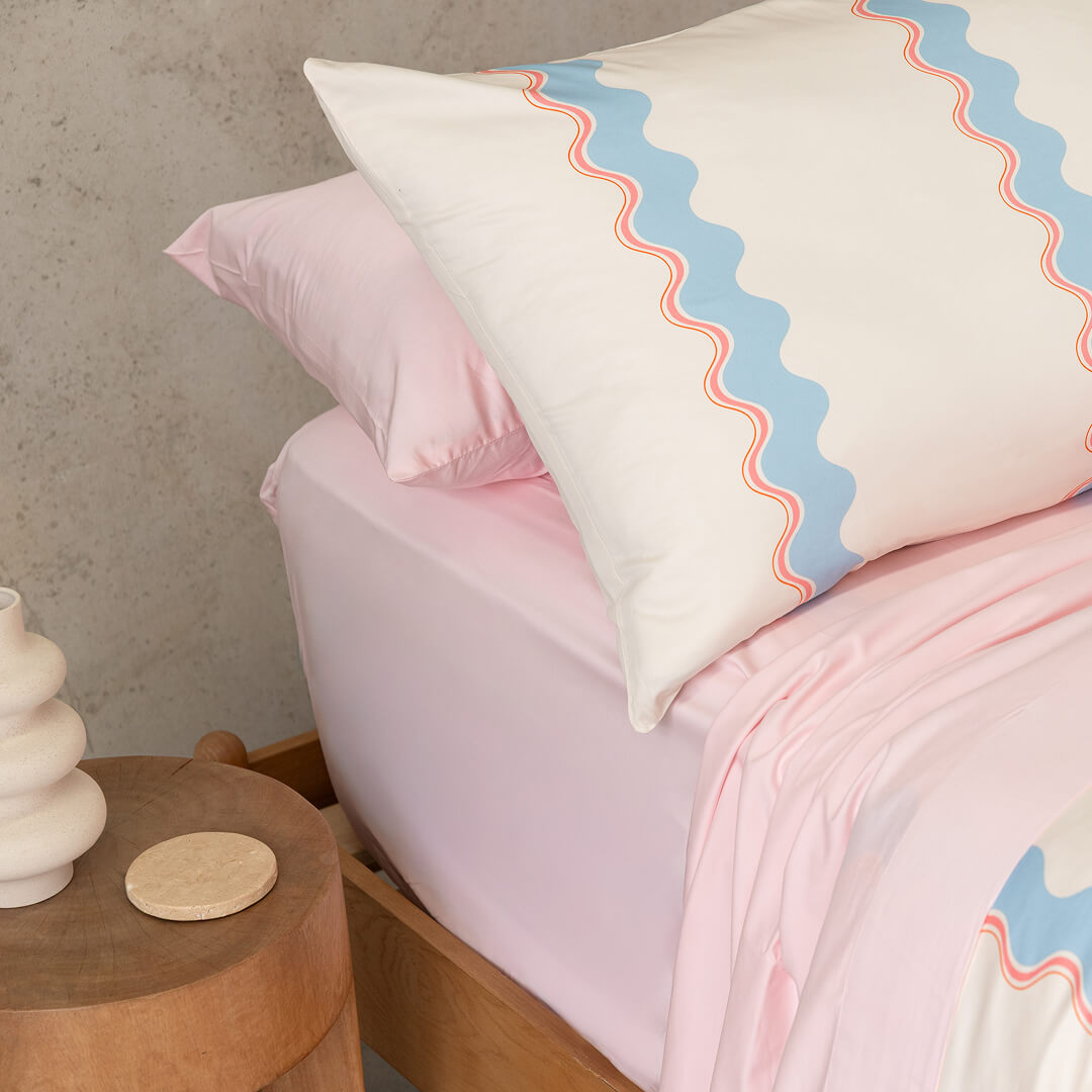 Bed with pink bedding and a decorative pillow with wavy pattern, wooden side table with white vase and coaster.