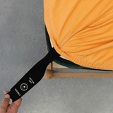 Hand pulling a black elastic corner band labeled 'Bottom Left' to secure a mustard yellow fitted sheet around a mattress corner