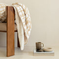 Wooden chair with a plaid blanket draped over it, next to a small table with a mug and books on a beige background.