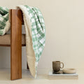 Green checkered blanket draped over a wooden chair with a cup and book on a table.