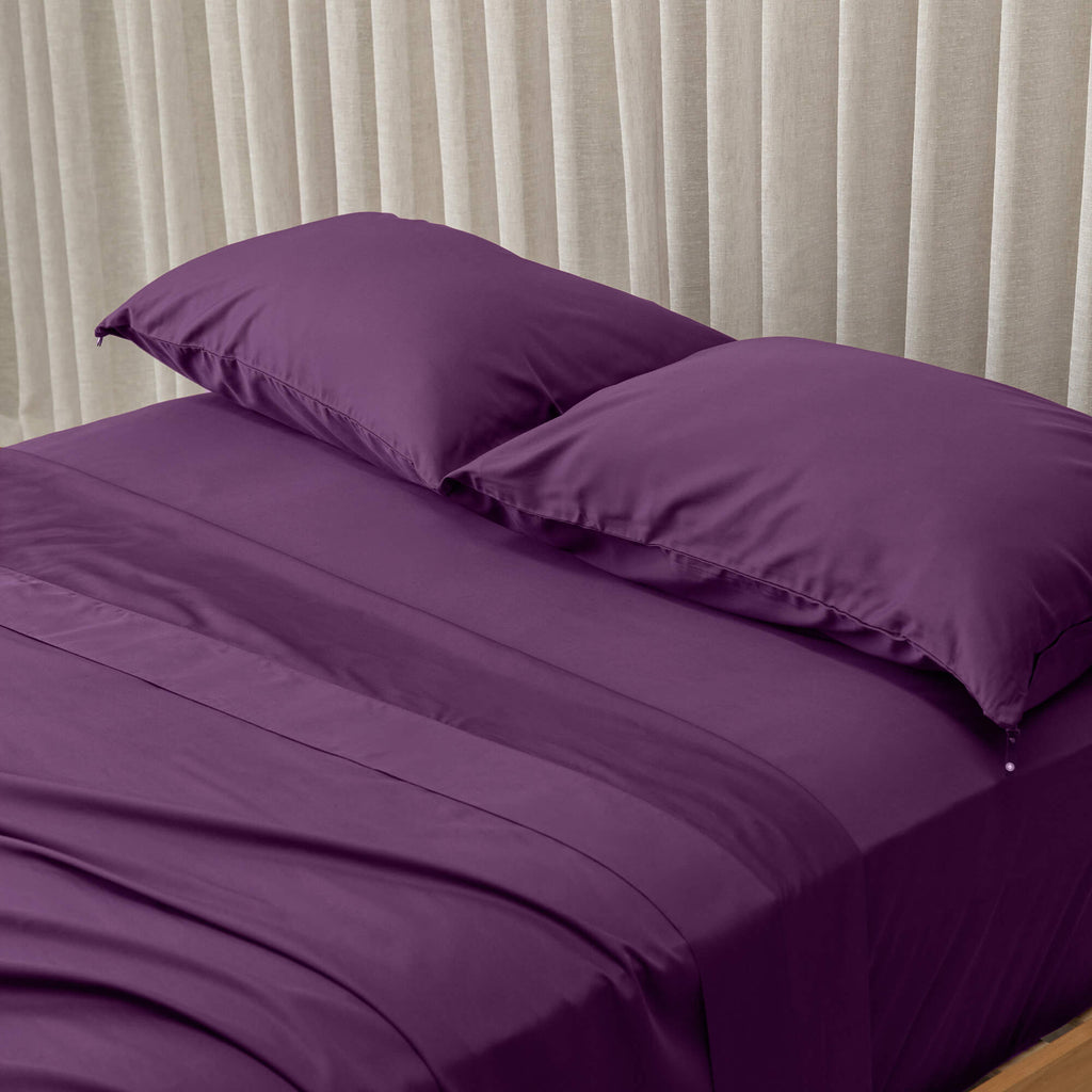 purple bedspread and pillows on a bed with beige curtains in the background