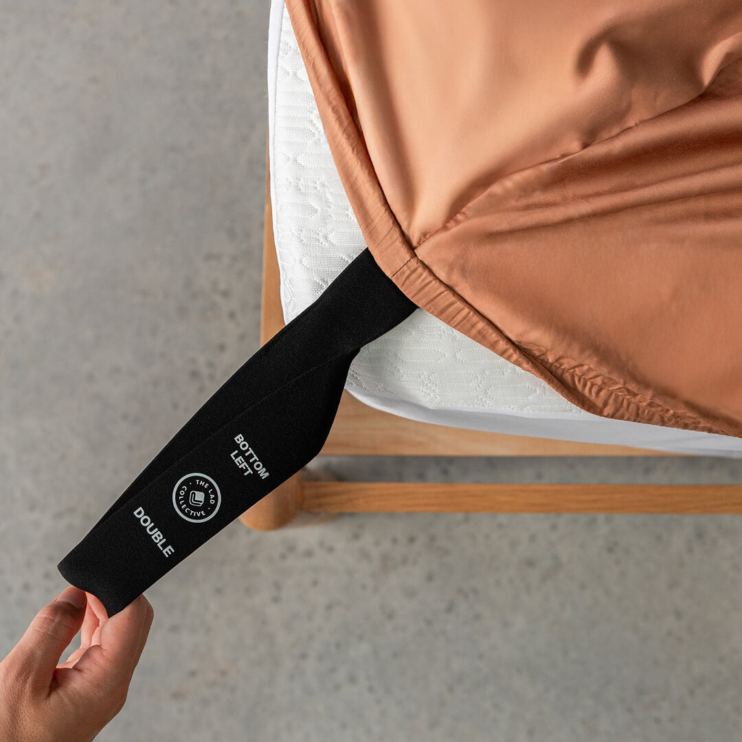 Close-up of a brown fitted sheet with a black strap being pulled by a hand onto the bed.