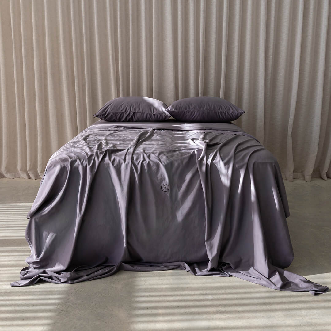 Bed with grey bedding and pillows against a beige curtain background.