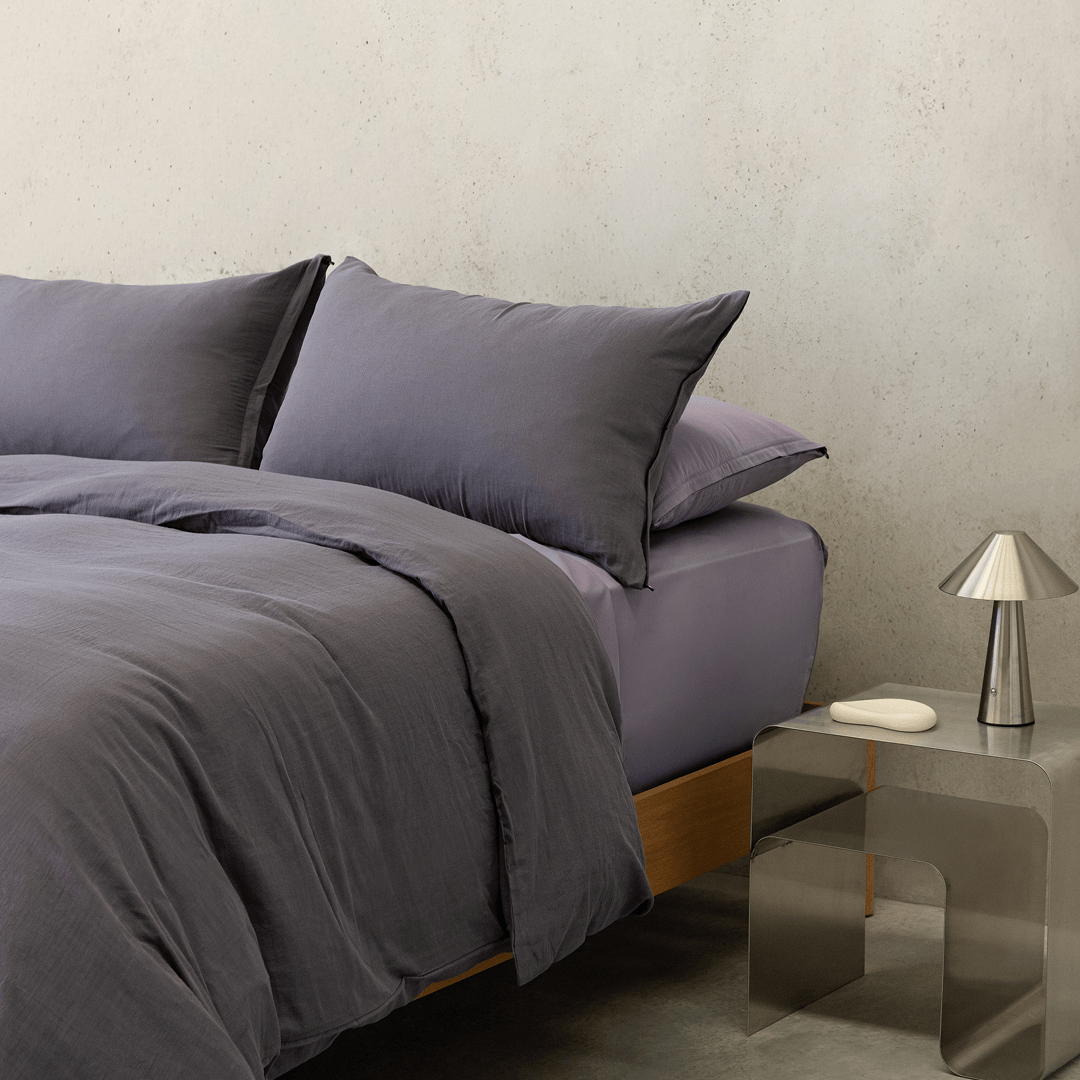 Styled dark stone gray bedding with matching pillows beside a metallic bedside table and silver lamp.